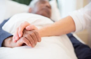 A person gently holds the hand of an elderly man lying in a hospital bed, conveying care and support.