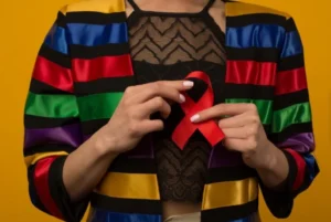 A woman wearing a colorful striped jacket holds a red ribbon, a universal symbol for HIV/AIDS awareness and support within the LGBTQ+ community, against a vibrant yellow background.