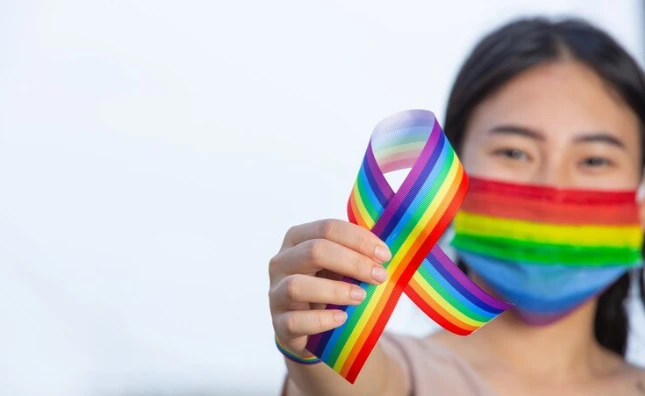 A young woman wearing a rainbow face mask holds up a rainbow ribbon, symbolizing LGBTQ+ pride and HIV awareness, advocating for inclusivity and health within the community, with a blurred white background.