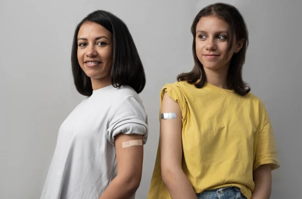Two smiling women, one in a white shirt and the other in a yellow shirt, display their bandaged arms after receiving vaccinations, promoting health, HIV prevention, and immunization awareness within the LGBTQ+ community.