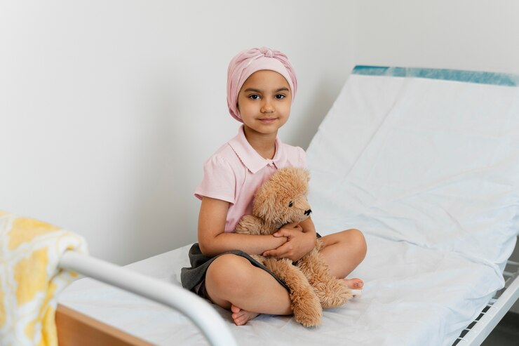 A child with a headscarf sitting on a hospital bed, hugging a teddy bear and smiling, representing resilience and hope in the fight against cancer.